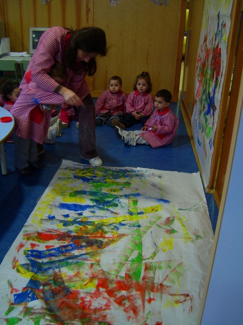 Niño ayudado por su profesora a pintar un mural, mientras otros niños sentados lo miran.