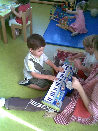 Niño tocando un teclado, mientras la profesora da palmas y otro niño mira un libro.