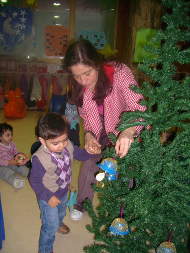 Niño ayudada por su porfesora a colocar adorno de navidad en un árbol.