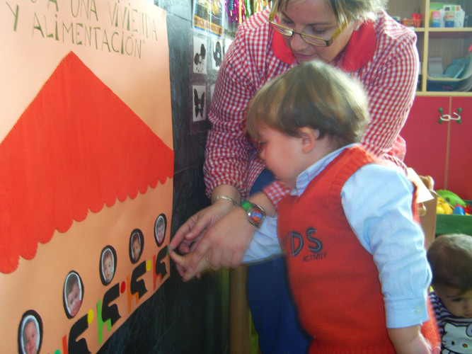 Niño ayudado por su profesora a pegar un punto verde en un cartel sobre un monigote que tiene su foto.