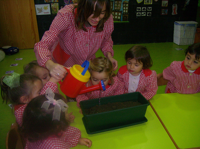 Niños junto a su profesora, regando una maceta de tierra.