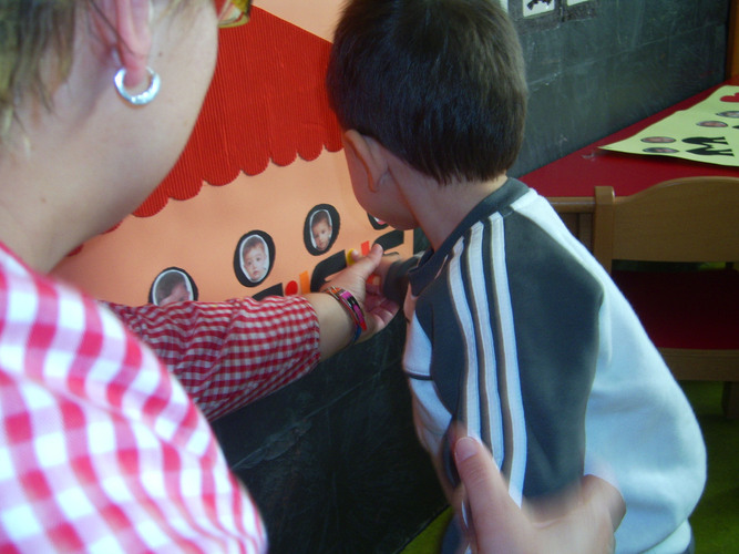 Niño ayudado por su profesora a pegar un punto amarillo en un cartel sobre un monigote que tiene su foto.