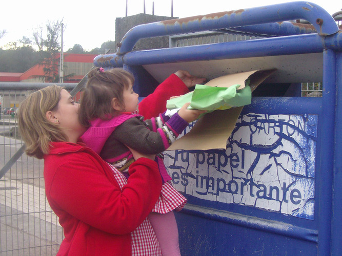 Profesora ayudando a una niña a introducir cartones en un contenedor de reciclaje.