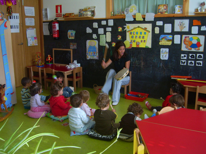 Grupo de niños sentados tocando sonajeros mientras la profesora toca la pandereta.