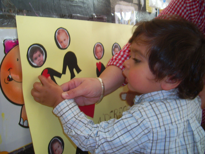 Niño ayudado por su profesora a pegar un punto verde en un cartel sobre un monigote que tiene su foto.