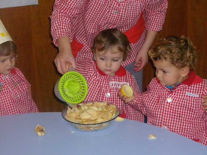 Profesora echando zumo de limón en el recipiente con trozos de manzana, mientras los niños la miran.