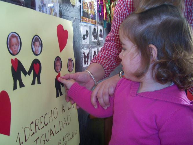 Niña ayudada por su profesora a pegar un corazón sobre un monigote con su foto, en un cartel.