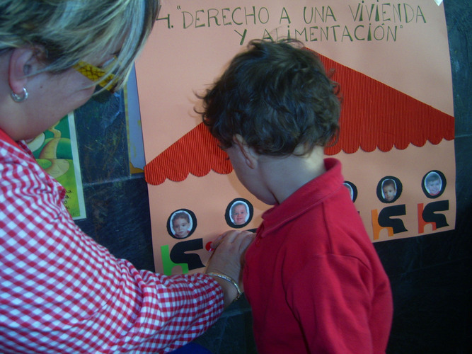 Niño ayudado por su profesora a pegar un punto rojo en un cartel sobre un monigote que tiene su foto.