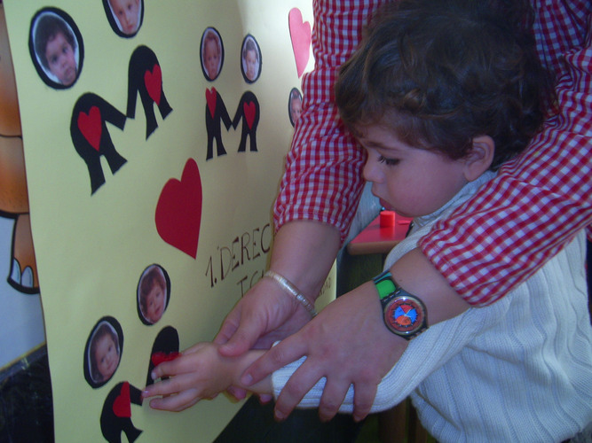 Niño ayudado por su profesora a pegar un corazón en un cartel sobre un monigote que tiene su foto.