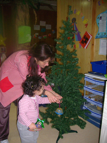 Niña ayudada por su porfesora a colocar adorno de navidad en un árbol.