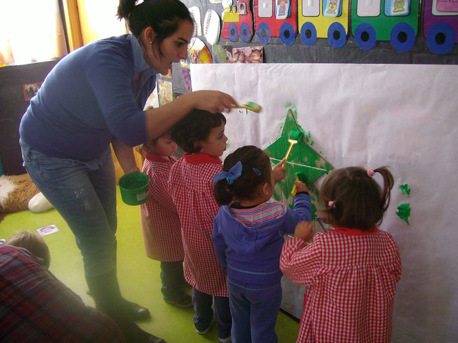 Niñas y su profesora pintando de verde un arbol de cartón pegado en un mural.