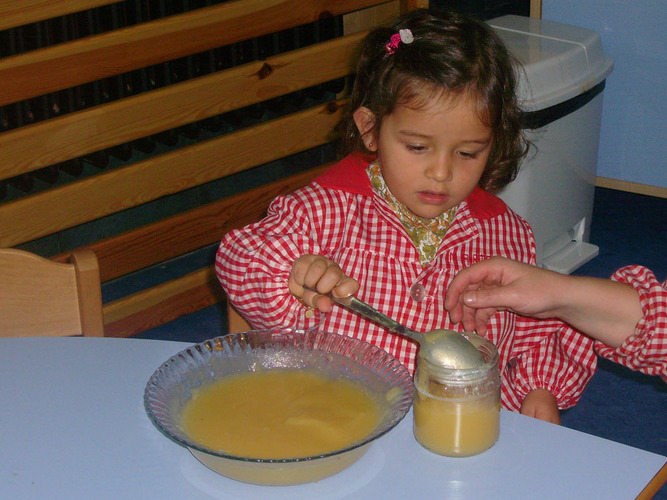 Niña echando la papilla del recipiente a un bote, mediante una cuchara.