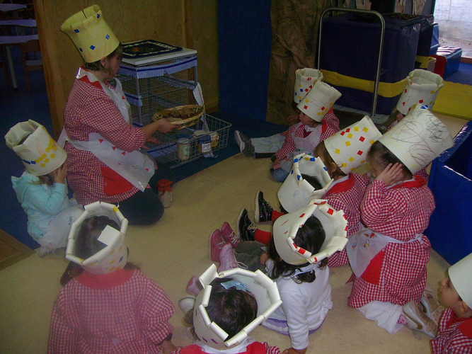 Profesora guardando las papillas y los frutos en un carrito despensa, mientras los niños con gorros de cocinero, la miran.