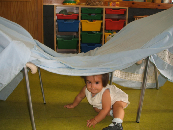 Niña pasando por debajo de una sábana colocada entre dos sillas.