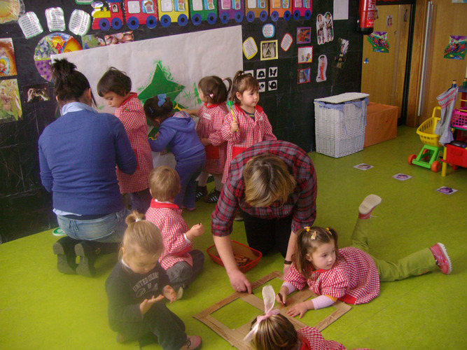 Niños ayudados por su profesora a pintar una ventana de cartón, para pegarla en el mural que pintan otros niños y su profesora, detrás.