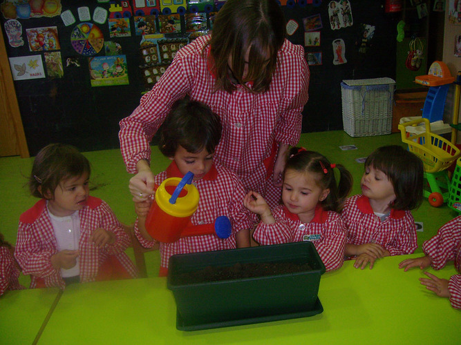 Niños junto a su profesora, regando una maceta de tierra.