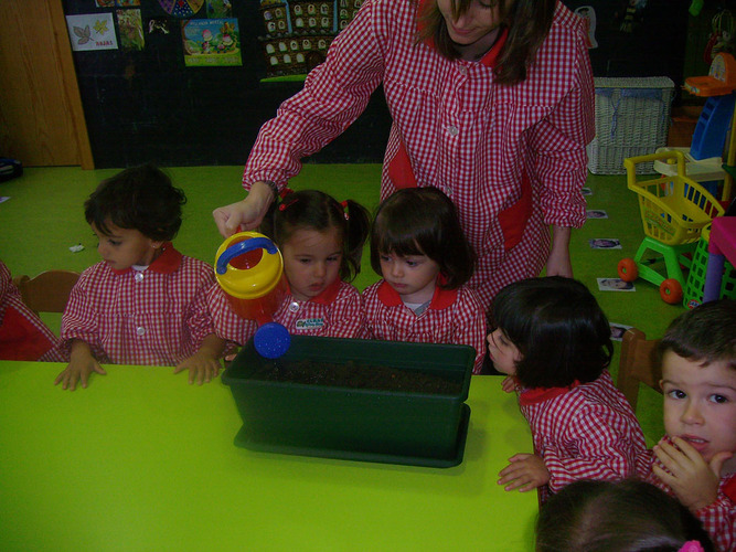 Niños junto a su profesora, regando una maceta de tierra.