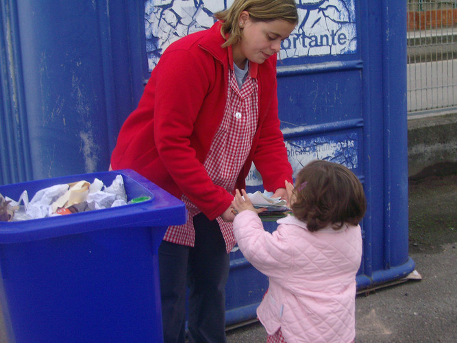 Profesora dandole papeles a una niña, para que ésta los recicle.