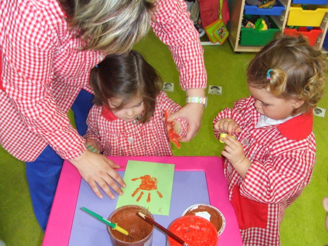 Profesora ayudando a una niña a pintar la palma de su mano en una hoja, mientas otra niña mira una pintura amarilla.